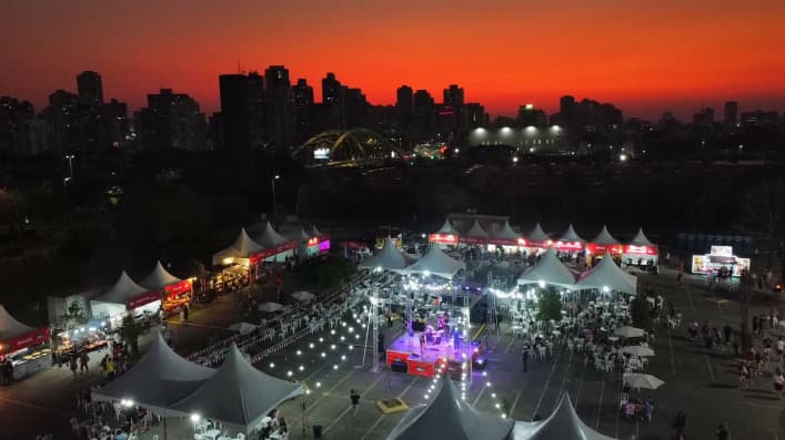 Imagem área externa de um evento, com palco e uma cidade com um por do sol ao fundo