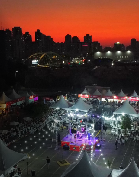 Imagem área externa de um evento, com palco e uma cidade com um por do sol ao fundo